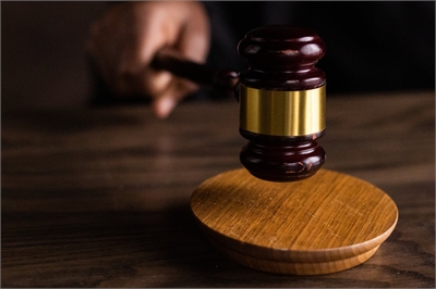 [ai] A close-up of a wooden gavel poised above a circular wooden sound block, with a blurred hand in the background, suggesting a courtroom setting.